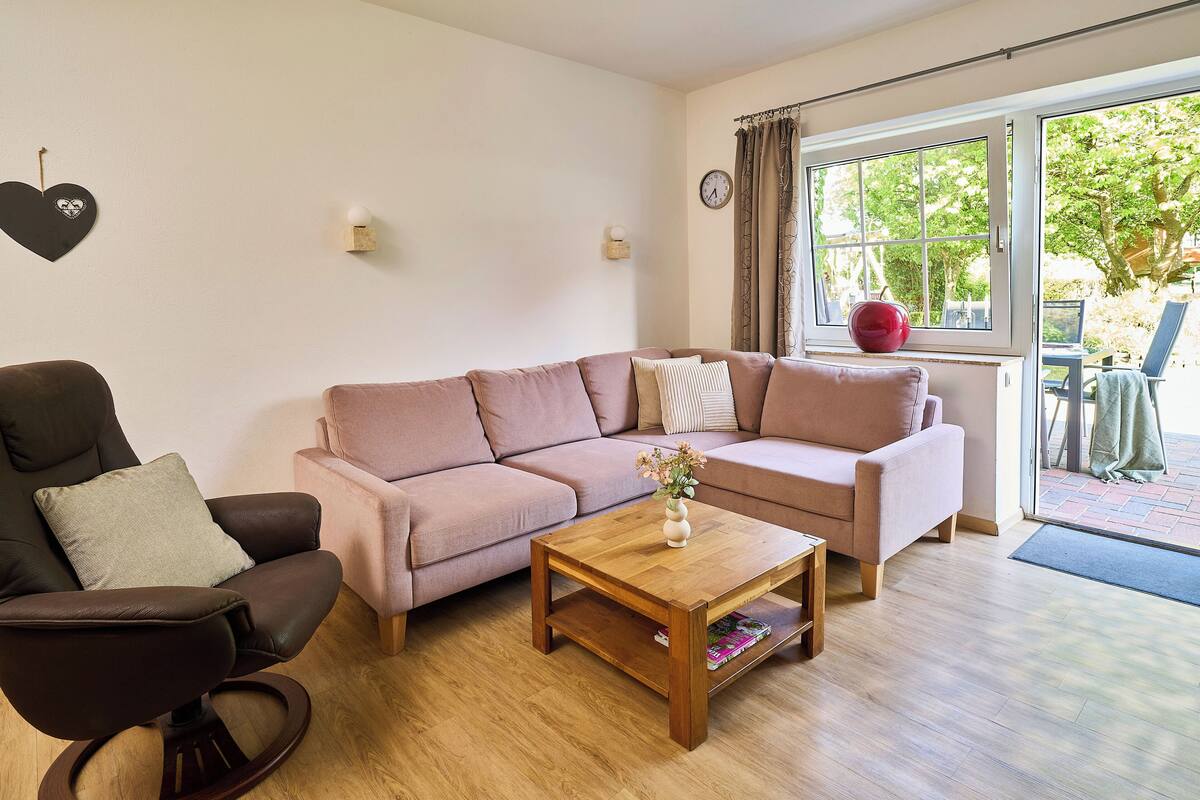 A living room with a sofa, armchair, and coffee table. There is a window with curtains and a view of the outdoors.