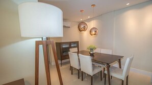 A dining area with a wooden table, white chairs, a lamp, and pendant lights. - PFino 206 - Apto Alto Padrão (Bombinhas)