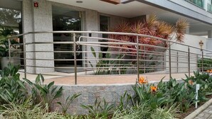 A modern building entrance with a metal railing, stone steps, and various plants including flowering ones. - PFino 206 - Apto Alto Padrão (Bombinhas)