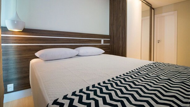 A modern hotel room with a large bed, a wooden headboard, a nightstand with a lamp, and a wardrobe. - PFino 206 - Apto Alto Padrão (Bombinhas)