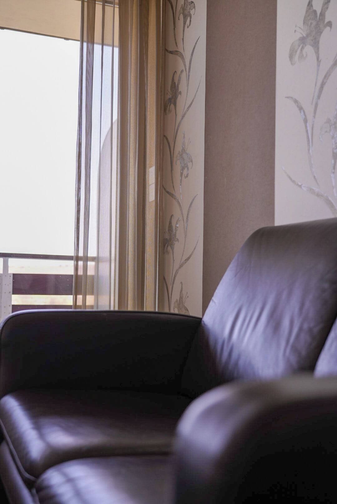 A leather chair in a room with a window, a wall with floral wallpaper, and a wooden railing.