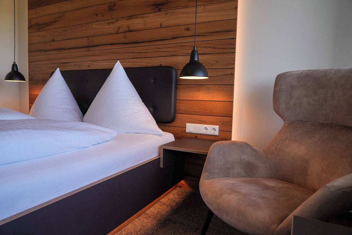 Une chambre d’hôtel moderne avec un mur en bois, un lit recouvert de draps blancs, un fauteuil marron et une petite table de chevet avec une lampe.