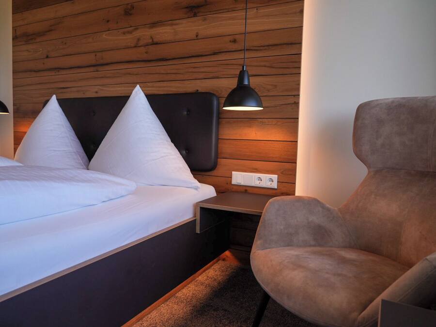 A modern hotel room with a wooden wall, a bed with white linens, a brown armchair, and a small bedside table with a lamp.