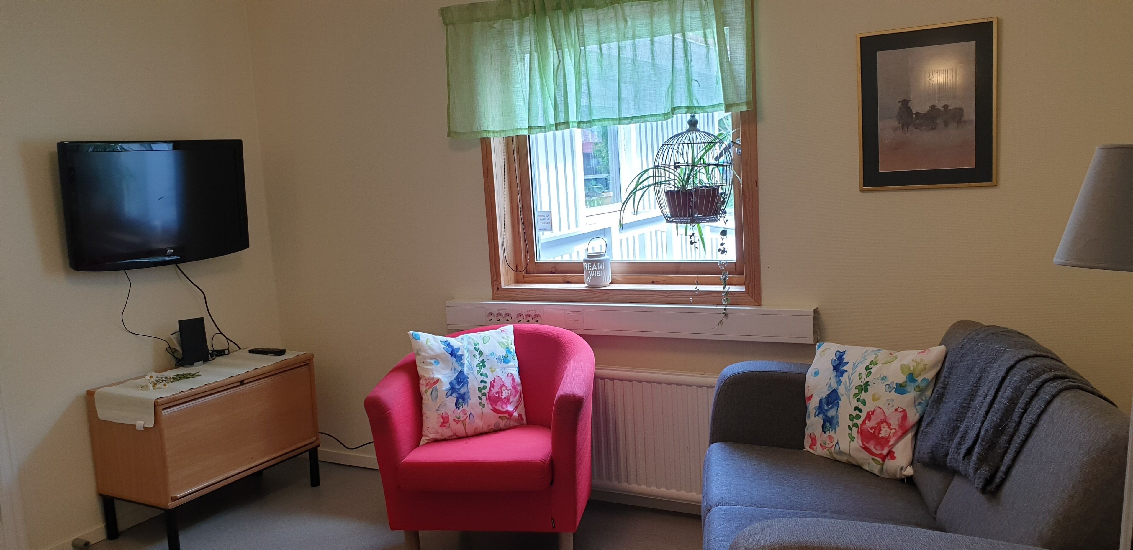 Ein Wohnzimmer mit Flachbildfernseher an der Wand, einem Fenster mit grüner Vorhang, einem Heizkörper, einem Holznachttisch, einem roten Sessel und einem grauen Sofa mit blumigen Kissen.