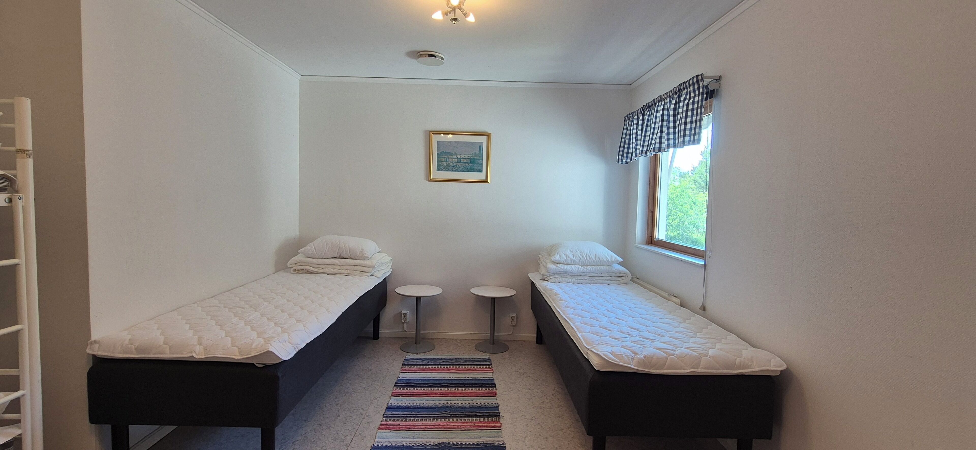 A small, clean room with two single beds, a small table, a chair, and a window with a view of greenery.
