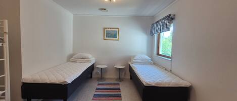 A small, clean room with two single beds, a small table, a chair, and a window with a view of greenery.