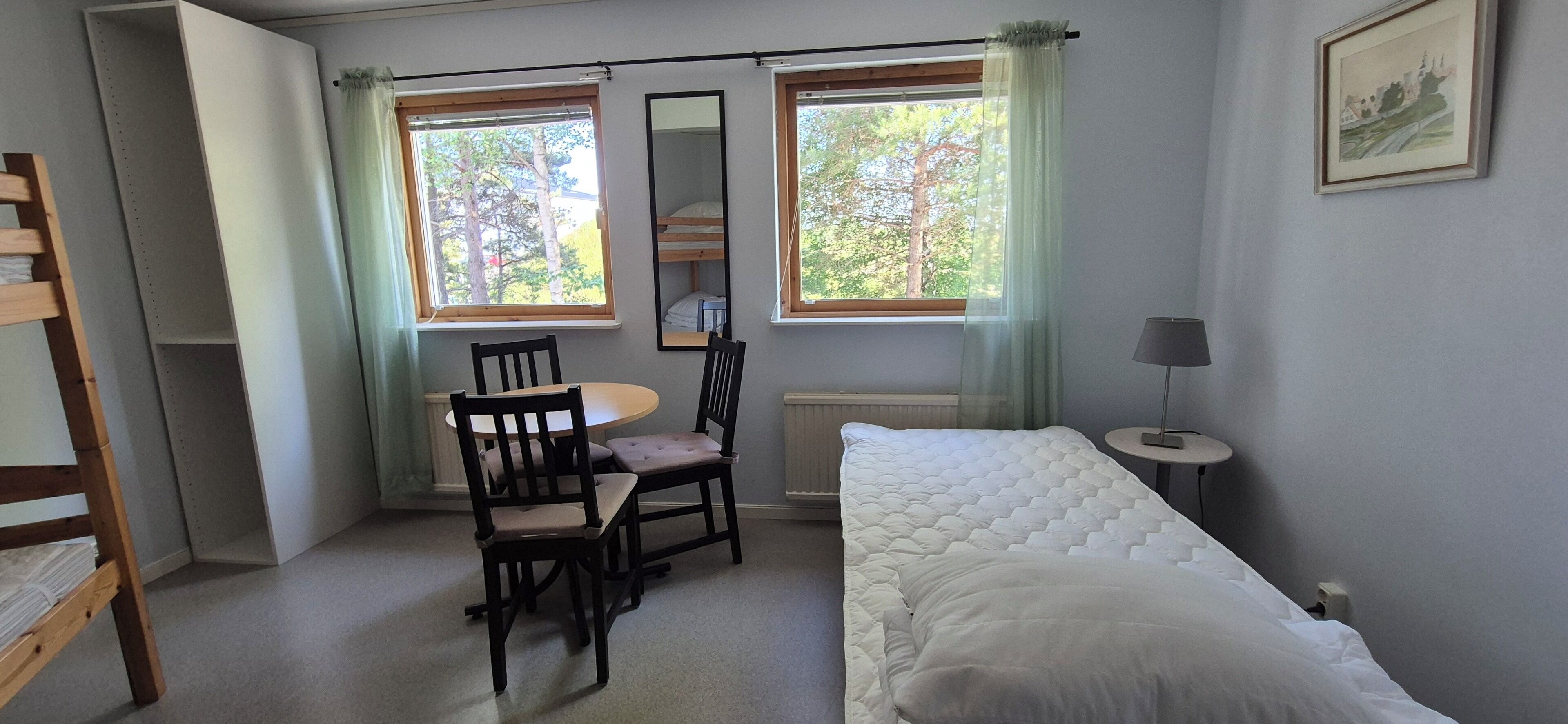 A room with a bed, a table with chairs, two windows with curtains, a radiator, and a framed picture on the wall.