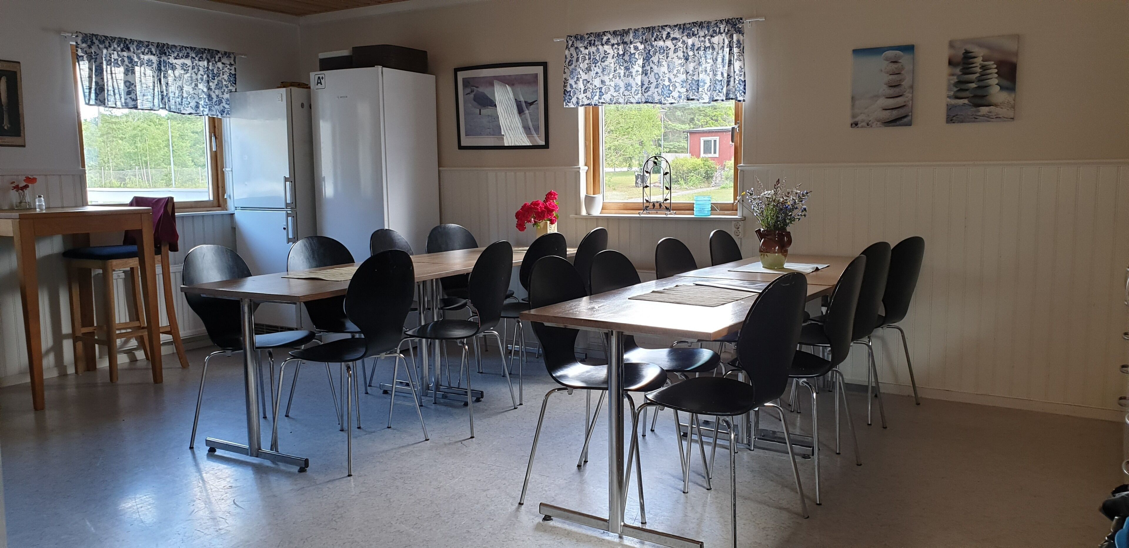 A dining area with a long table, black chairs, a wooden table, a refrigerator, and a window with a view of a house.