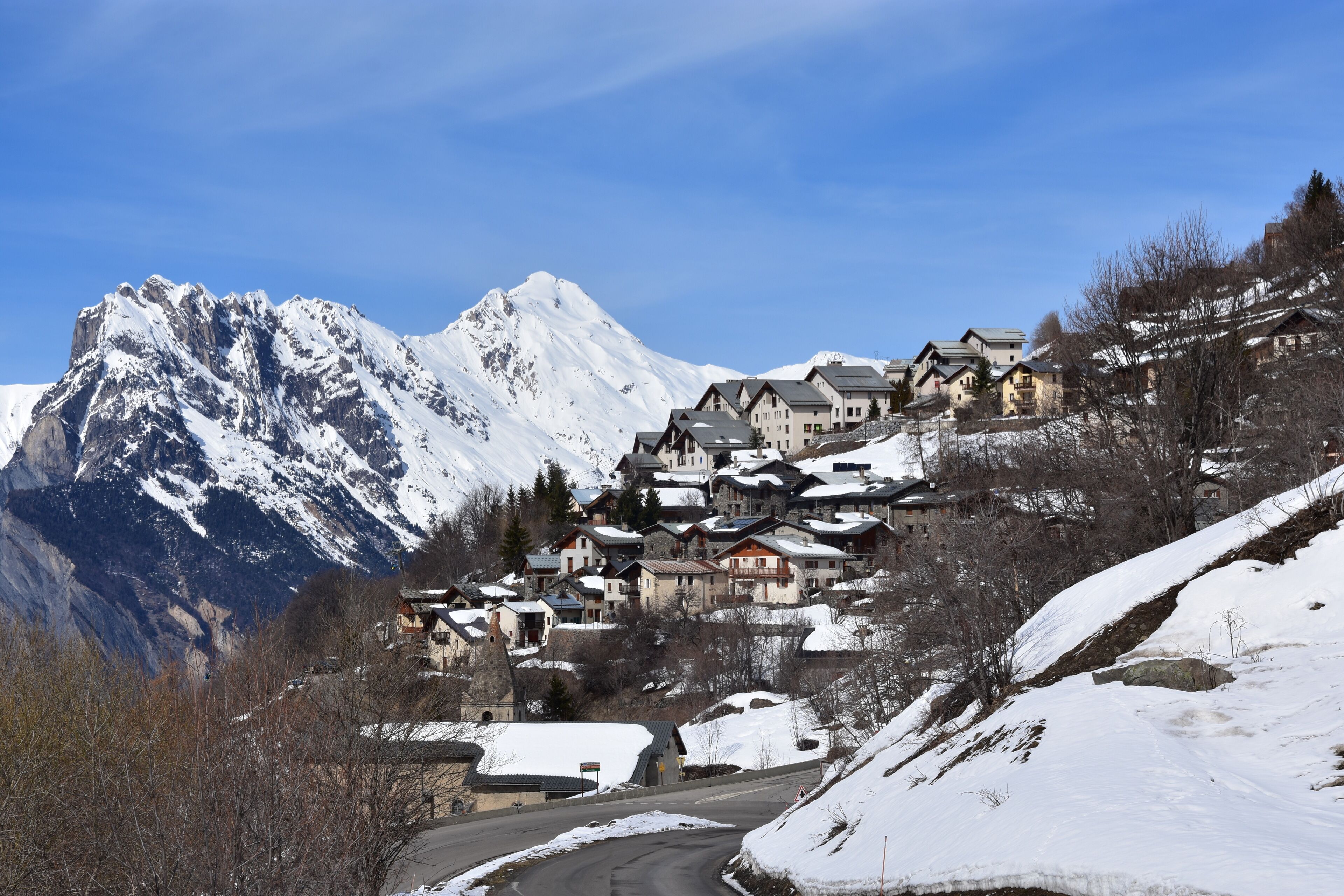 A snow-covered mountain village with traditional houses, a winding road, and a prominent mountain peak in the background.