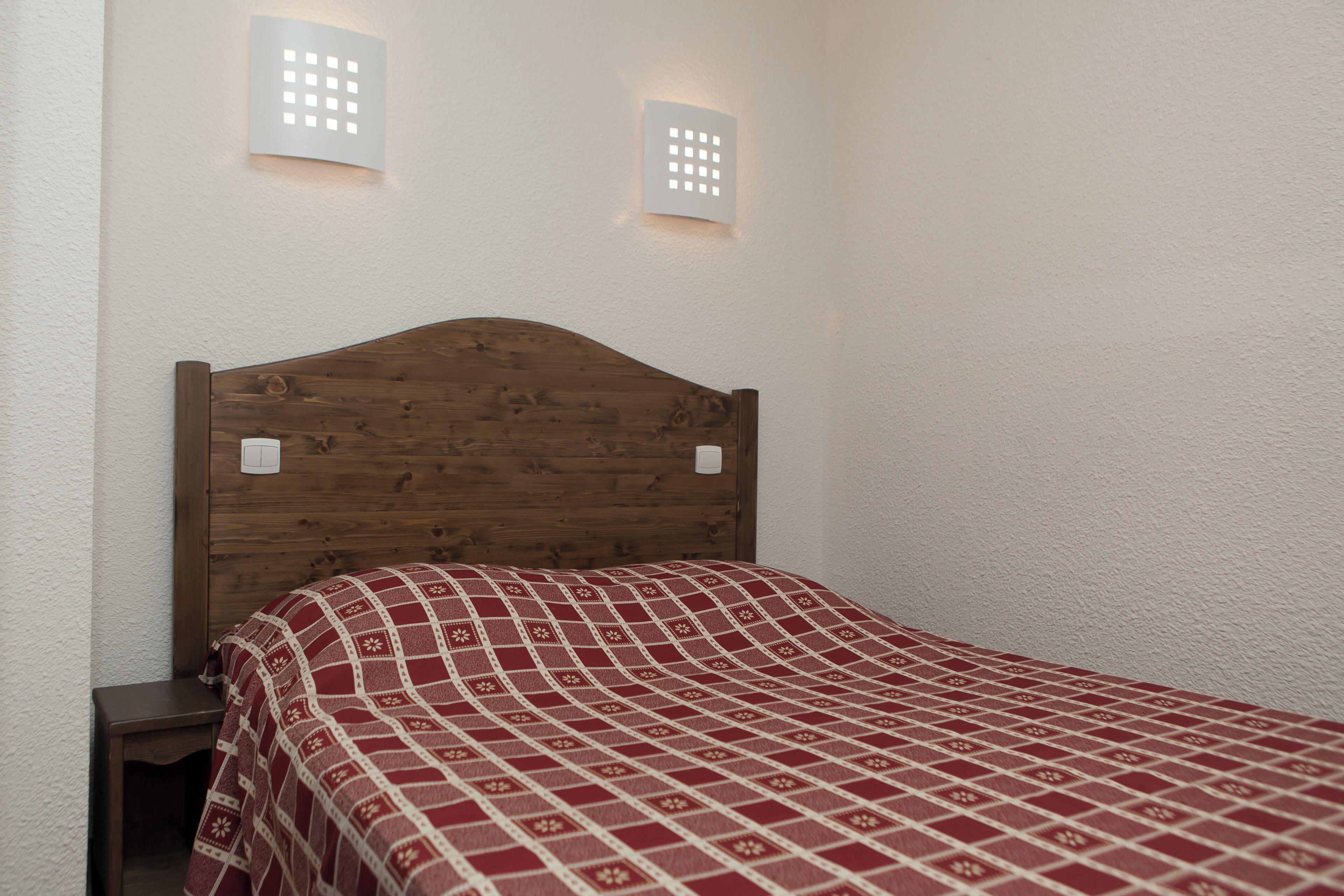 A single bed with a wooden headboard and a red patterned bedspread in a room with white walls and two wall-mounted light fixtures.