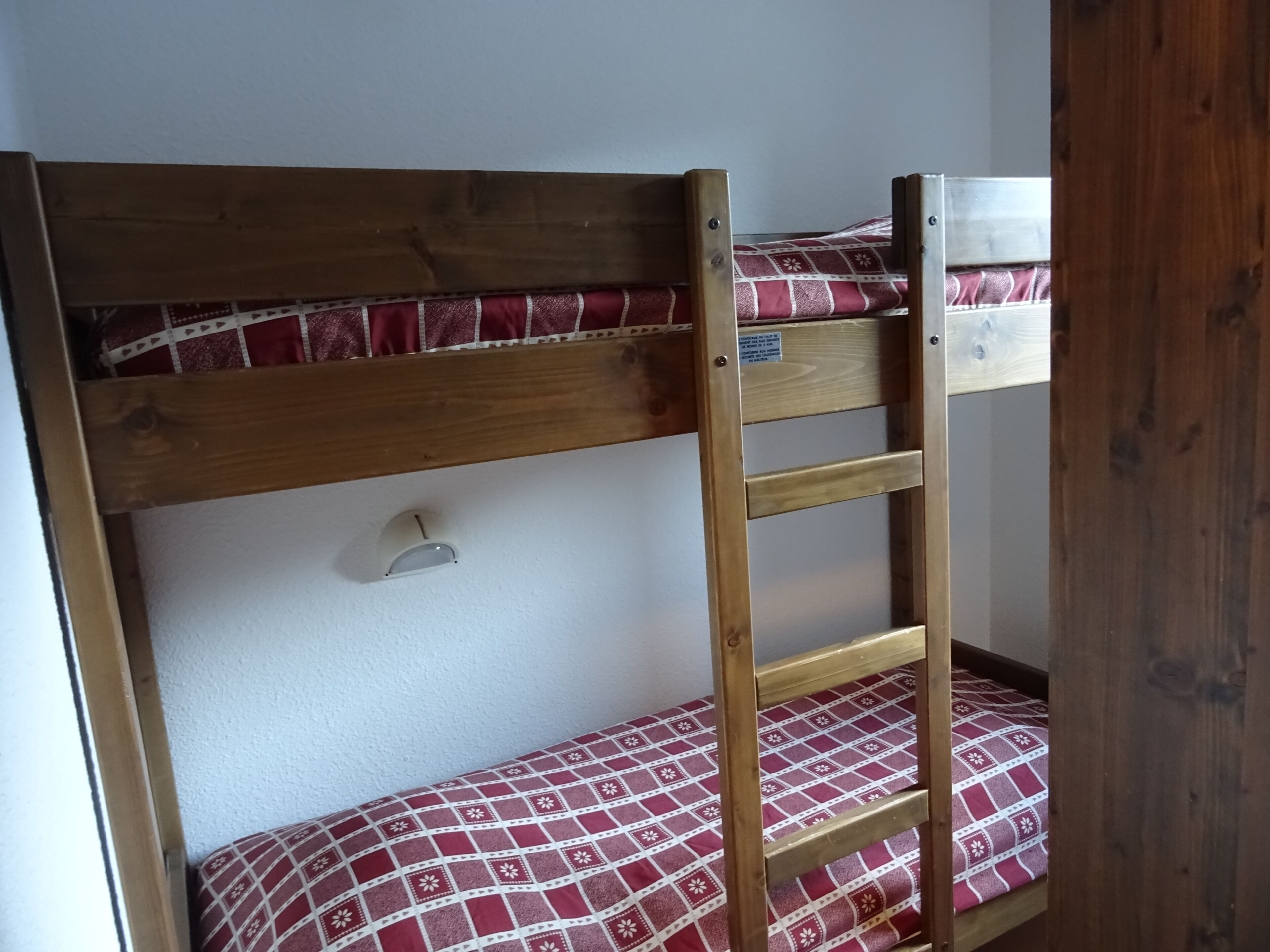 A bunk bed with a wooden frame, a mattress with a red and white pattern, and a small shelf with a light fixture.