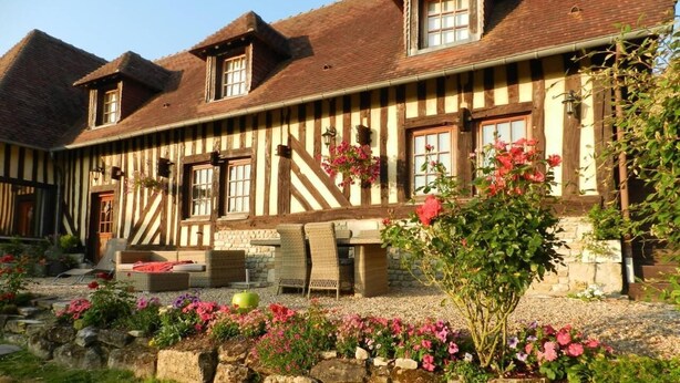 A traditional half-timbered building with a stone foundation, outdoor seating, and a garden with flowers. - Le Pressoir (Beuvron-en-Auge)