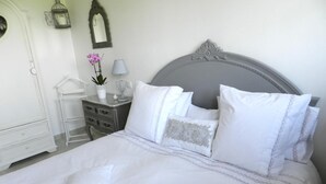 A neatly made bed with white bedding and multiple pillows, a bedside table with a lamp and a vase of flowers, and a dresser with a mirror and a small plant. - Le Pressoir (Beuvron-en-Auge)