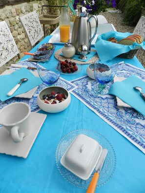 Mesa de pequeno-almoço posta com toalha azul, jarra de café, copo de sumo de laranja, tigela de fruta, chávena e prato com faca e colher.