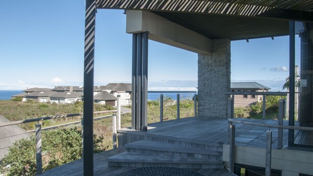 A modern outdoor deck with a stone pillar, a circular seating area, and a view of coastal buildings. - 10 Martingale (Knysna)