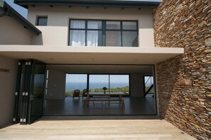 Una casa moderna con fachada de piedra, un amplio patio abierto con mesa y sillas, y vista al océano.