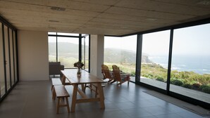 Une pièce spacieuse avec une grande table et des chaises, de grandes fenêtres offrant une vue sur l’océan et les collines.