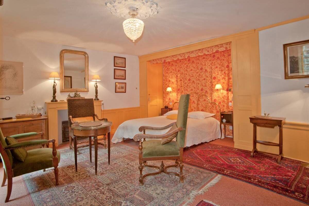 A traditional room with a bed, two chairs, a table, a chandelier, and a fireplace.