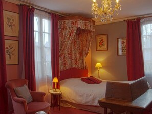 A bedroom with a bed, a chair, a table, a lamp, and a chandelier.