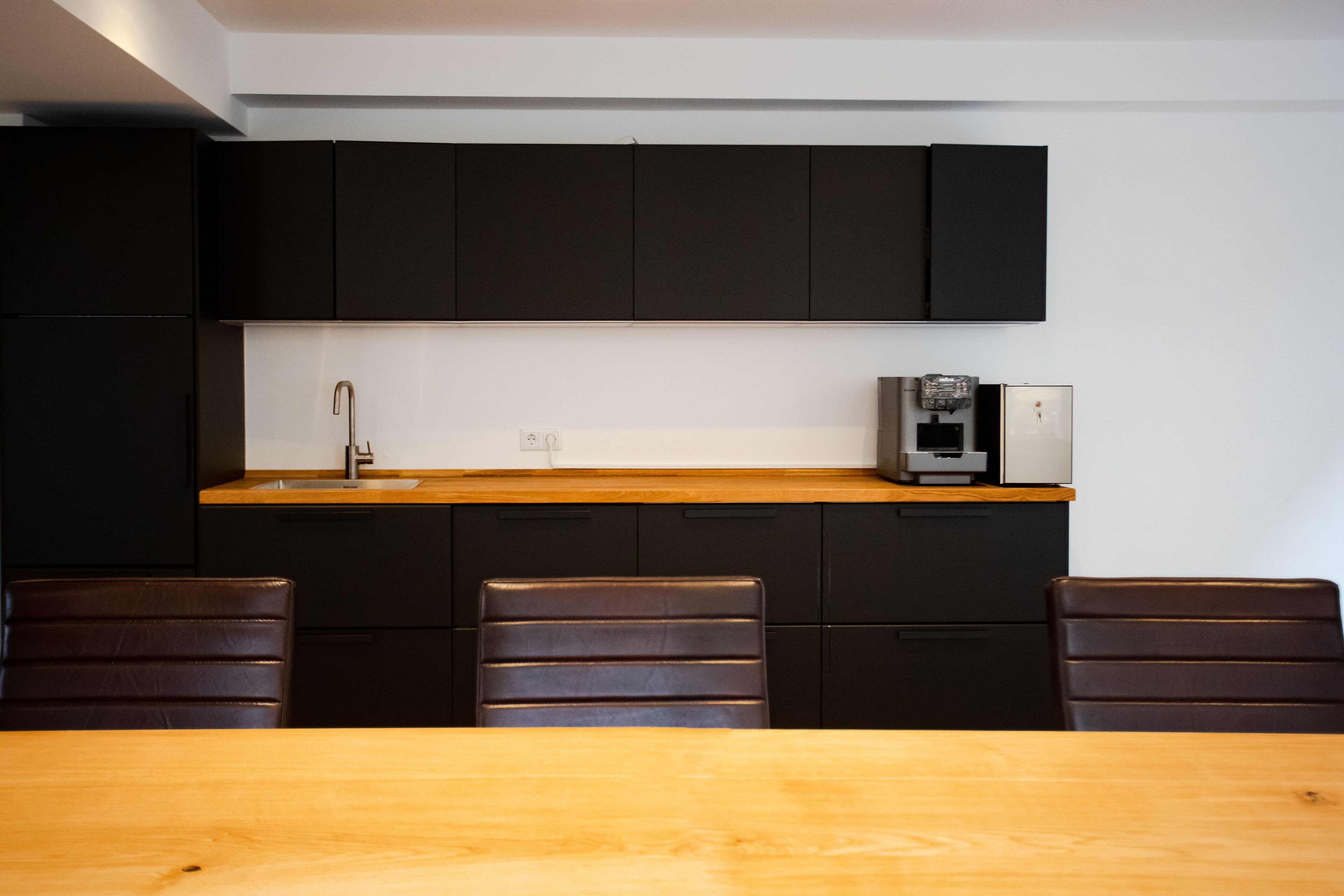 A modern kitchen with black cabinets, a wooden countertop, a sink, a coffee machine, and a dining area with a wooden table and chairs.