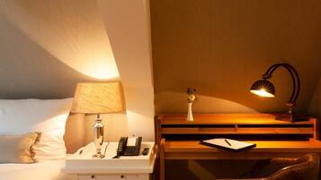 Habitación de hotel con cama, mesita de noche con lámpara, escritorio con lámpara, una silla y un pequeño jarrón con flores.