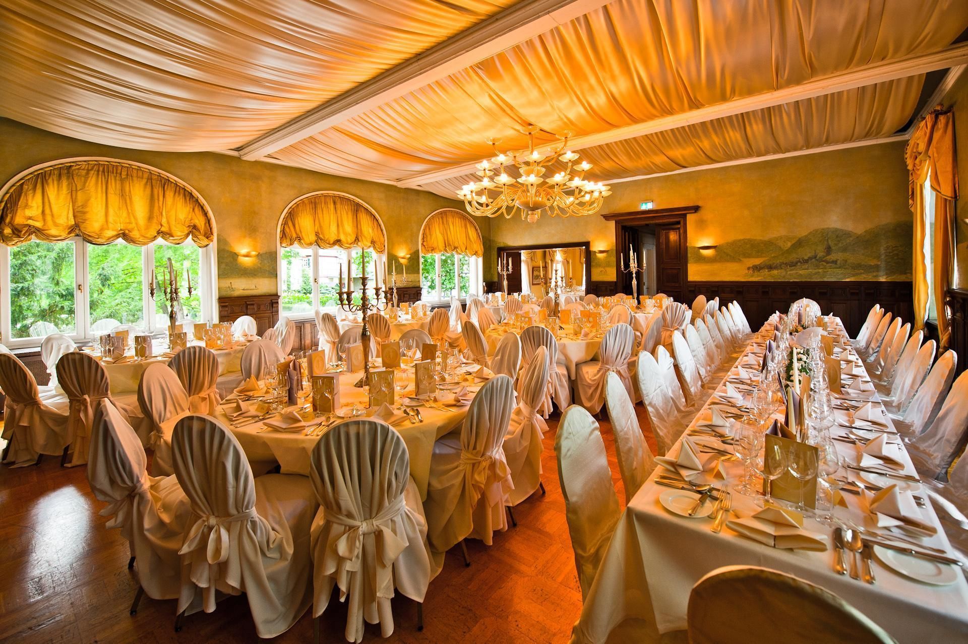 Un salón de banquetes con mesas redondas dispuestas para un evento formal, con candelabros y ventanas amplias con cortinas.