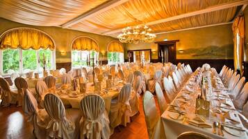 Un salón de banquetes con mesas redondas dispuestas para un evento formal, con candelabros y ventanas amplias con cortinas.