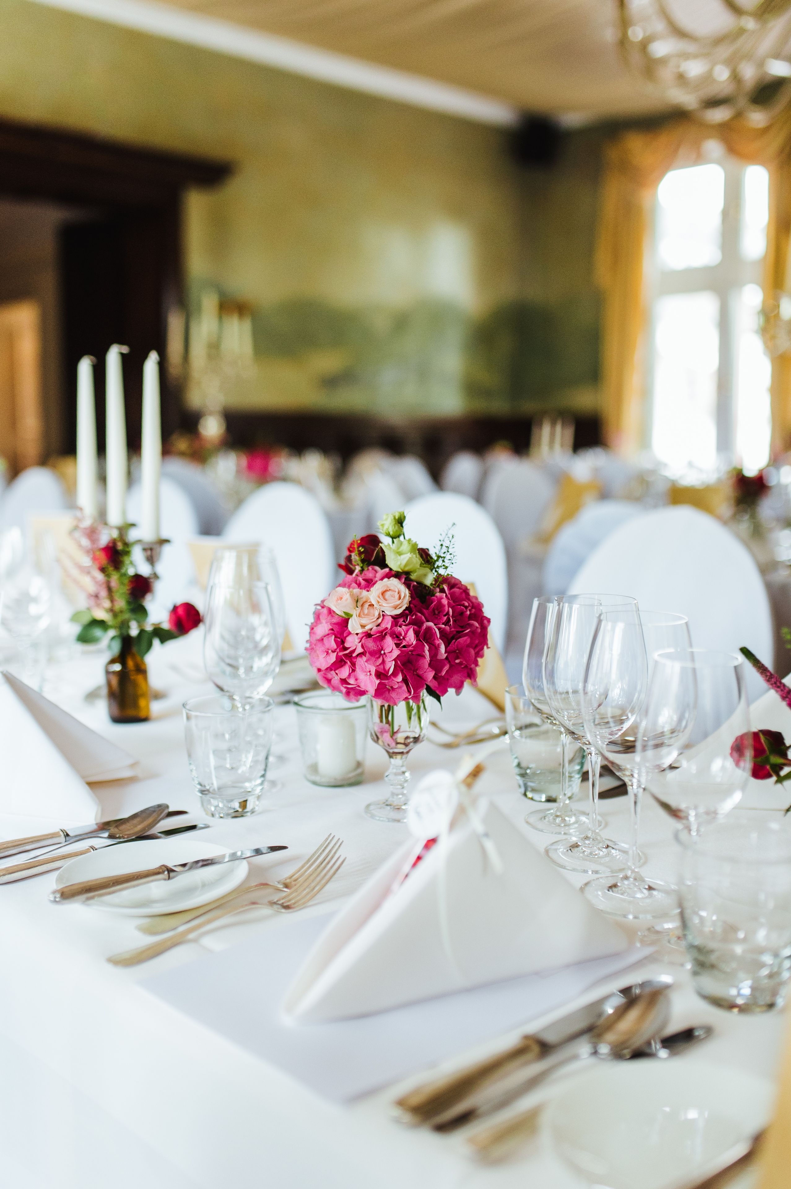 A table set for a formal event with white tablecloths, clear glassware, and pink floral centerpieces.