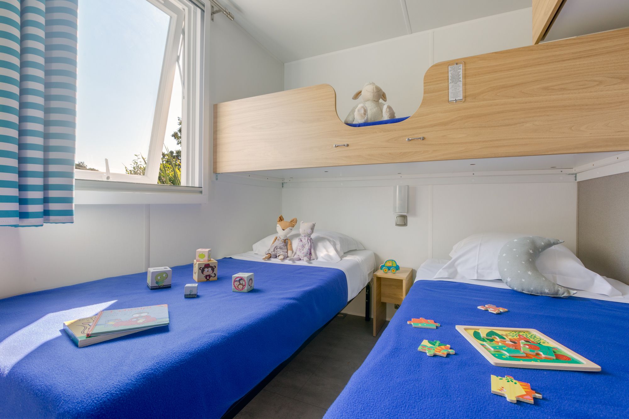 Een slaapkamer met twee eenpersoonsbedden, een loggia met gordijnen, een nachtkastje met lamp en een klein plankje met knuffeldiertjes.