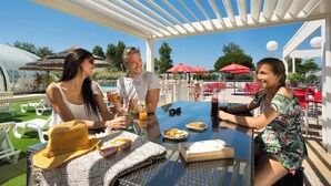 A covered outdoor dining area with a pool, red umbrellas, and white chairs. - Yelloh Village La Plage (Penmarch)