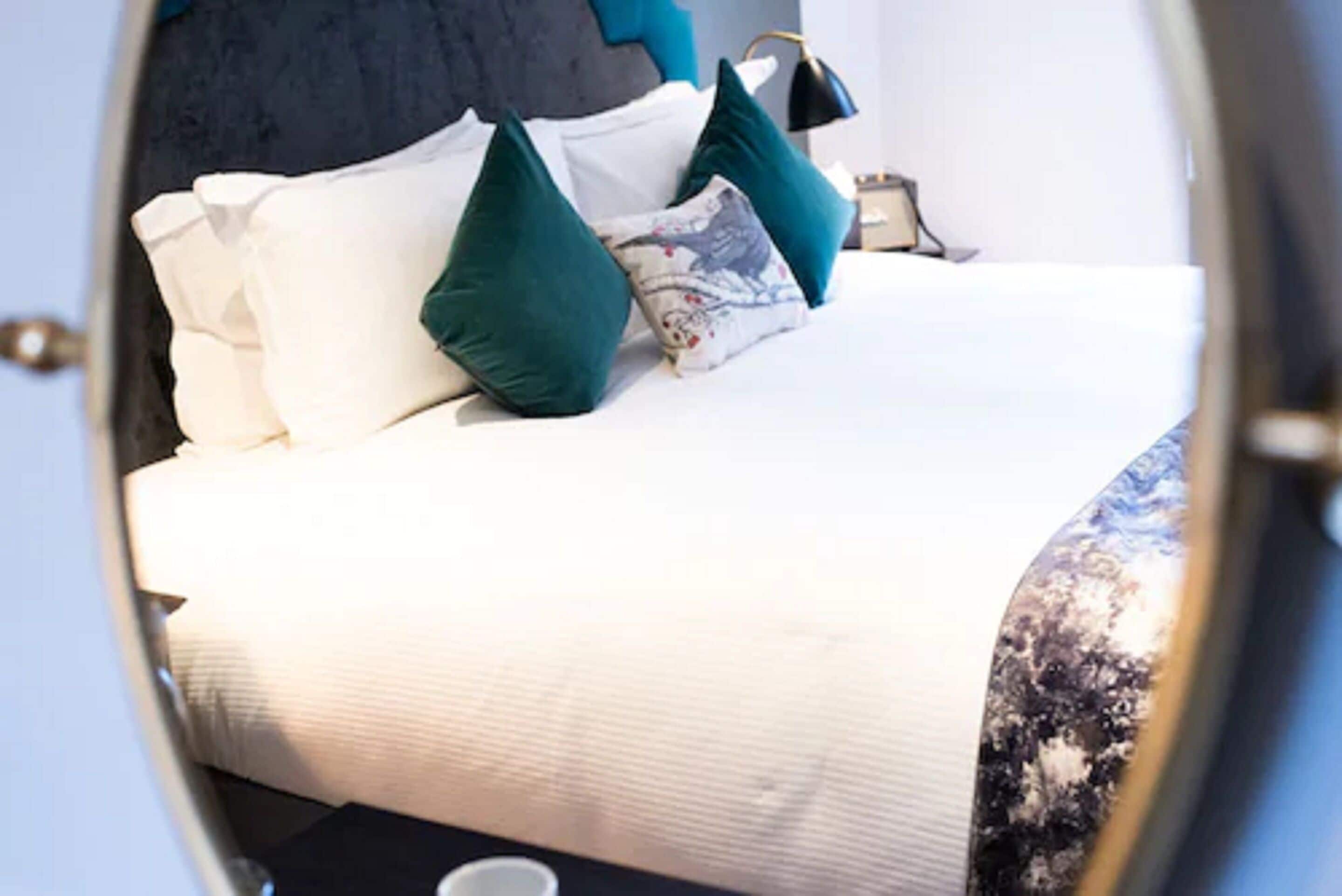 Una cama con ropa de cama blanca y almohadas color turquesa, una mesita de noche con una lámpara, y una ventana con vistas a árboles.