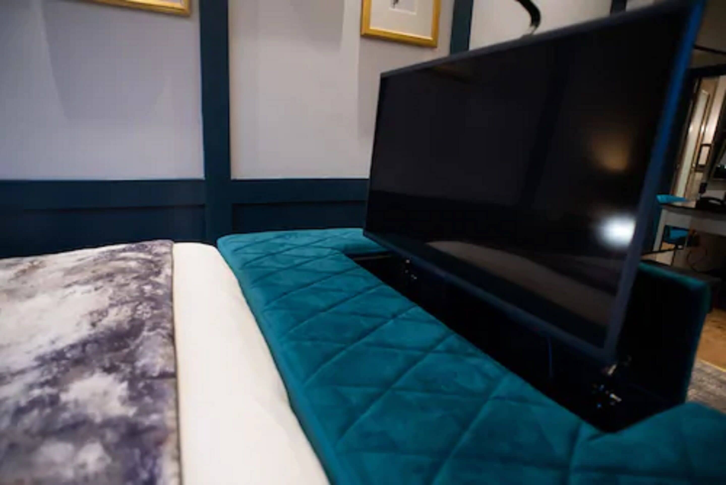 Una cama con cubrecama color aguamarina y almohada blanca, un televisor de pantalla plana sobre un soporte, y un cuadro en la pared.