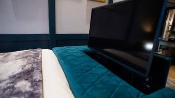 Una cama con cubrecama color aguamarina y almohada blanca, un televisor de pantalla plana sobre un soporte, y un cuadro en la pared.