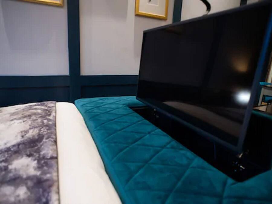 Una cama con cubrecama color aguamarina y almohada blanca, un televisor de pantalla plana sobre un soporte, y un cuadro en la pared.