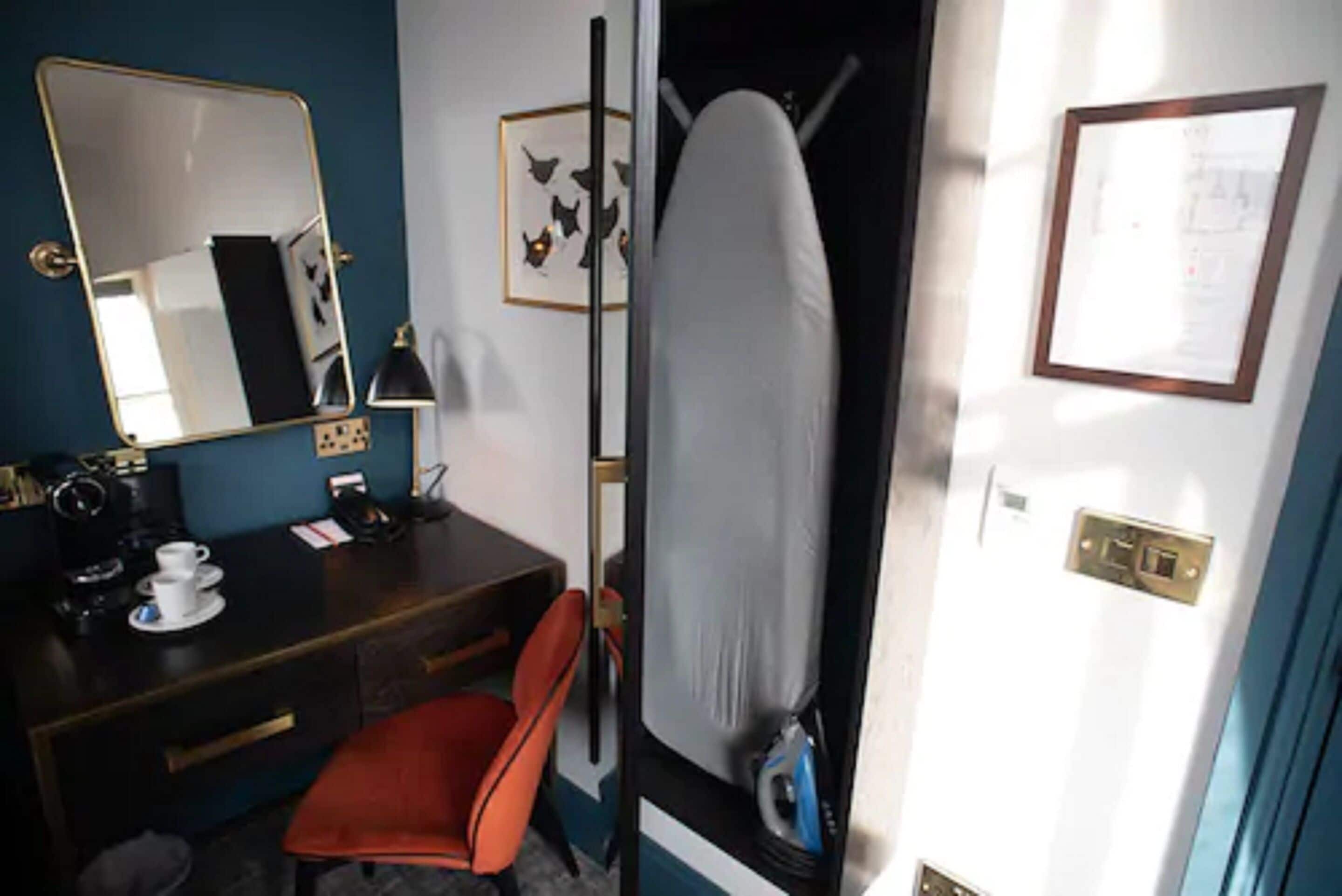 Habitación de hotel con escritorio, silla, espejo, tabla de planchar y una lámpara de pared.