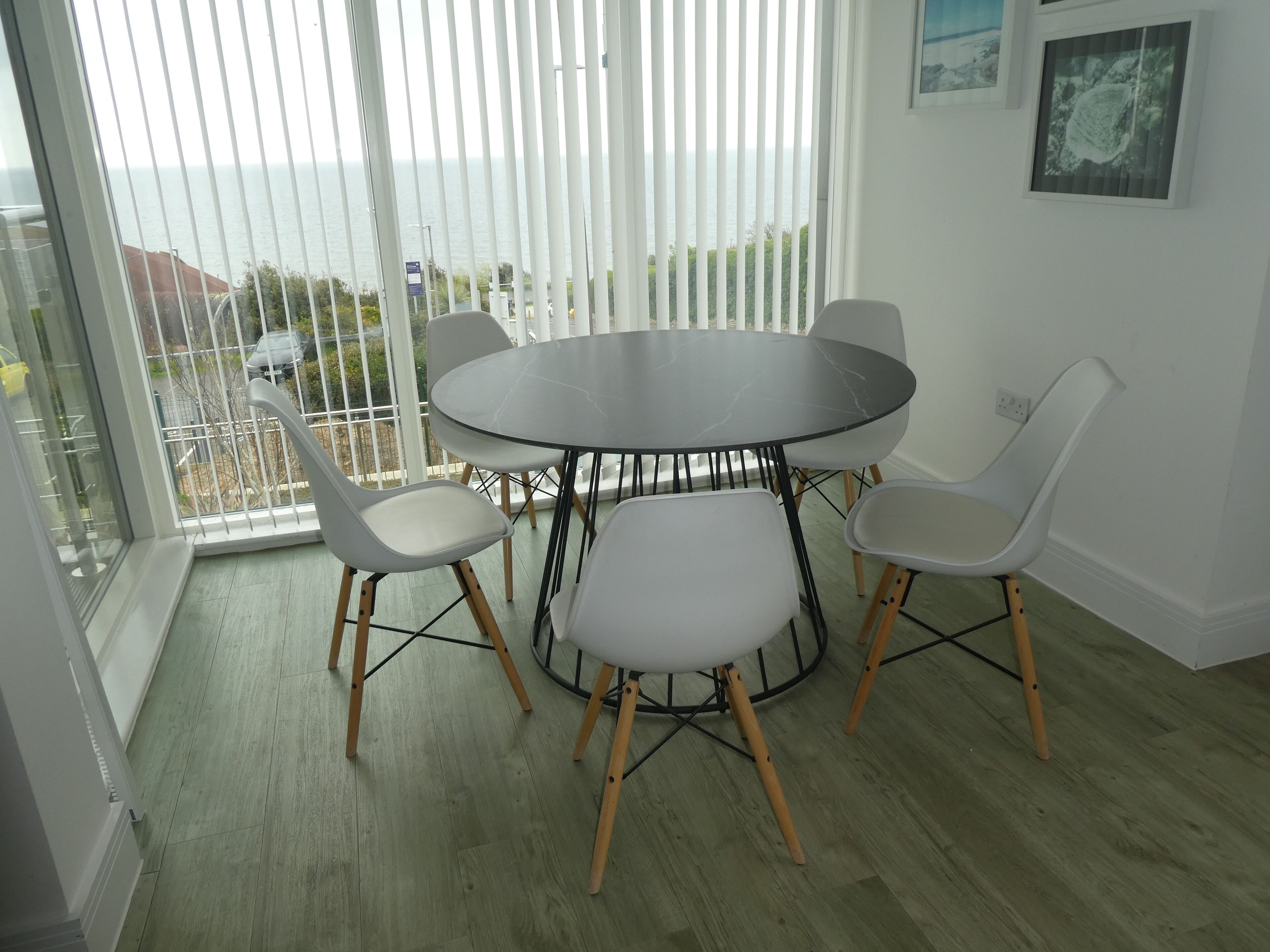 A dining area with a round table and six chairs, large windows with blinds, and a view of the sea.