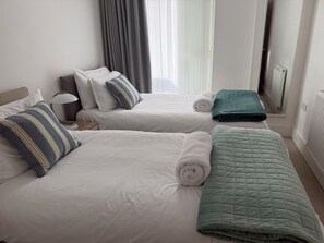 Habitación de hotel con dos camas, cada una con ropa blanca y una toalla enrollada. Una colcha verde acolchada cubre una de las camas. Una lámpara está sobre una mesita de noche, y se ve un radiador en la pared.