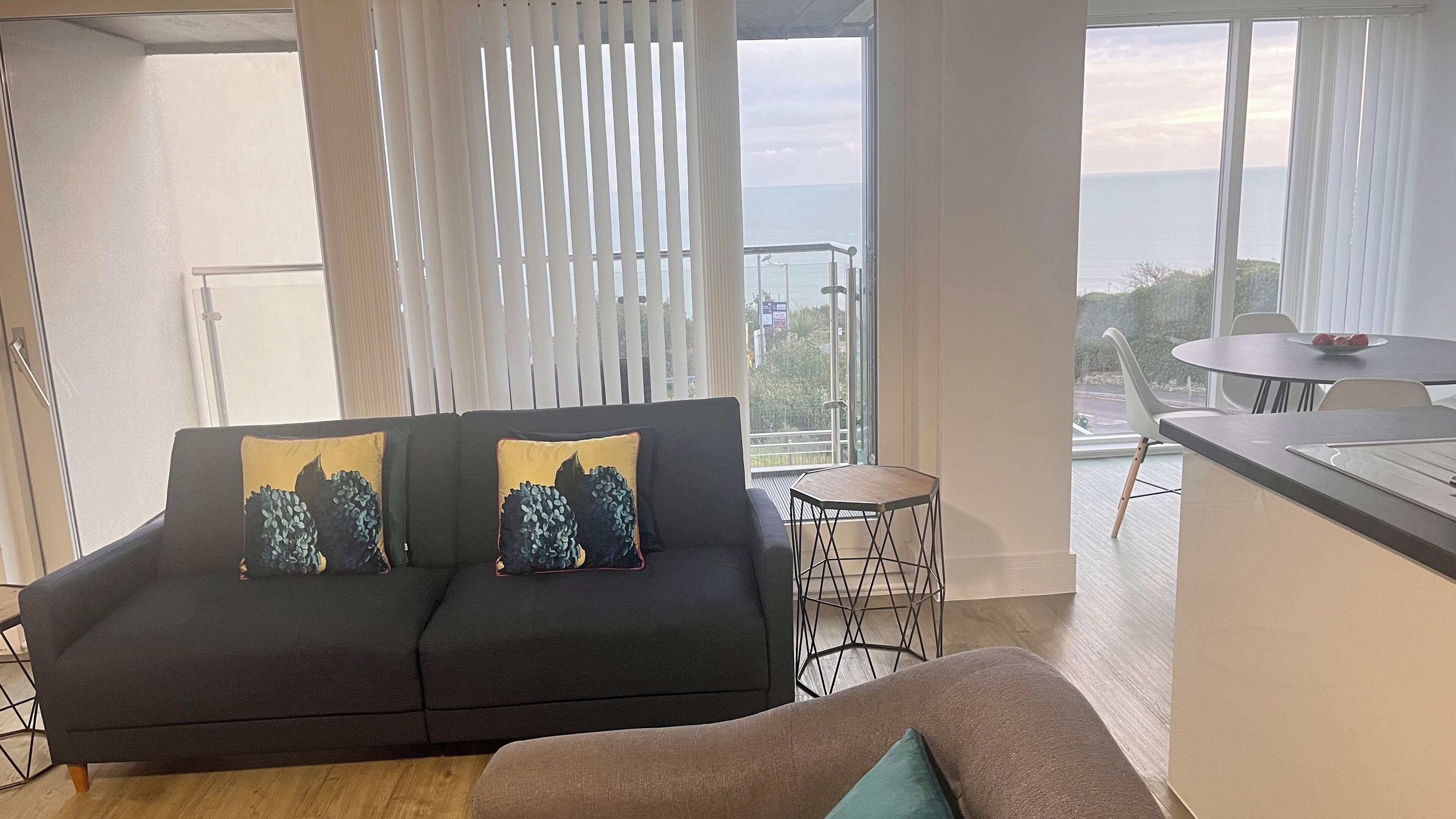Ein modernes Wohnzimmer mit einem dunkelgrauen Sofa, zwei dekorativen Kissen, einem kleinen runden Tisch und Meerblick durch große Fenster.