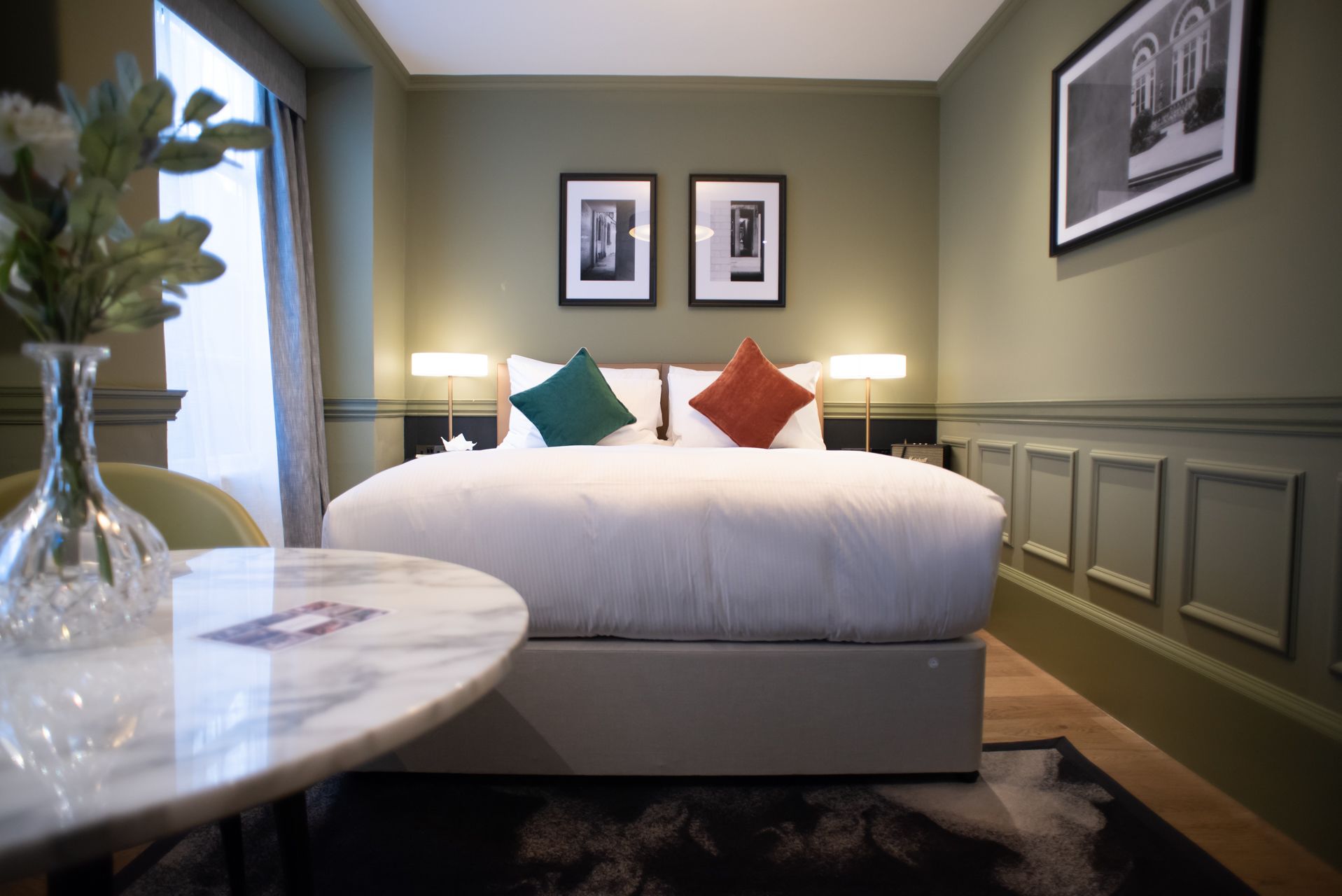 A hotel room with a large bed, two bedside lamps, a vase with flowers, and framed pictures on the wall.