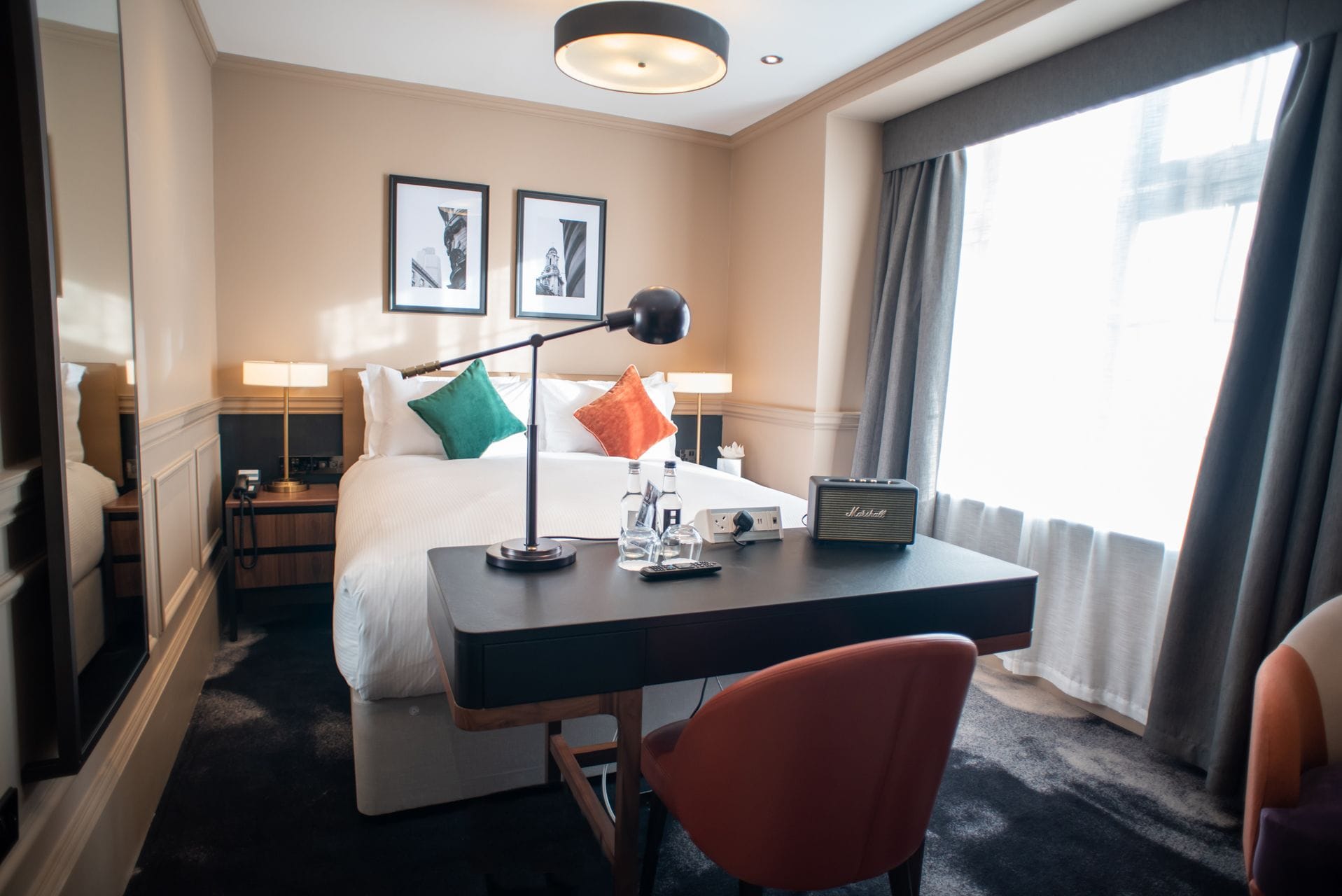 A hotel room with a bed, bedside tables, a desk, a chair, a lamp, a radio, and a window with curtains.