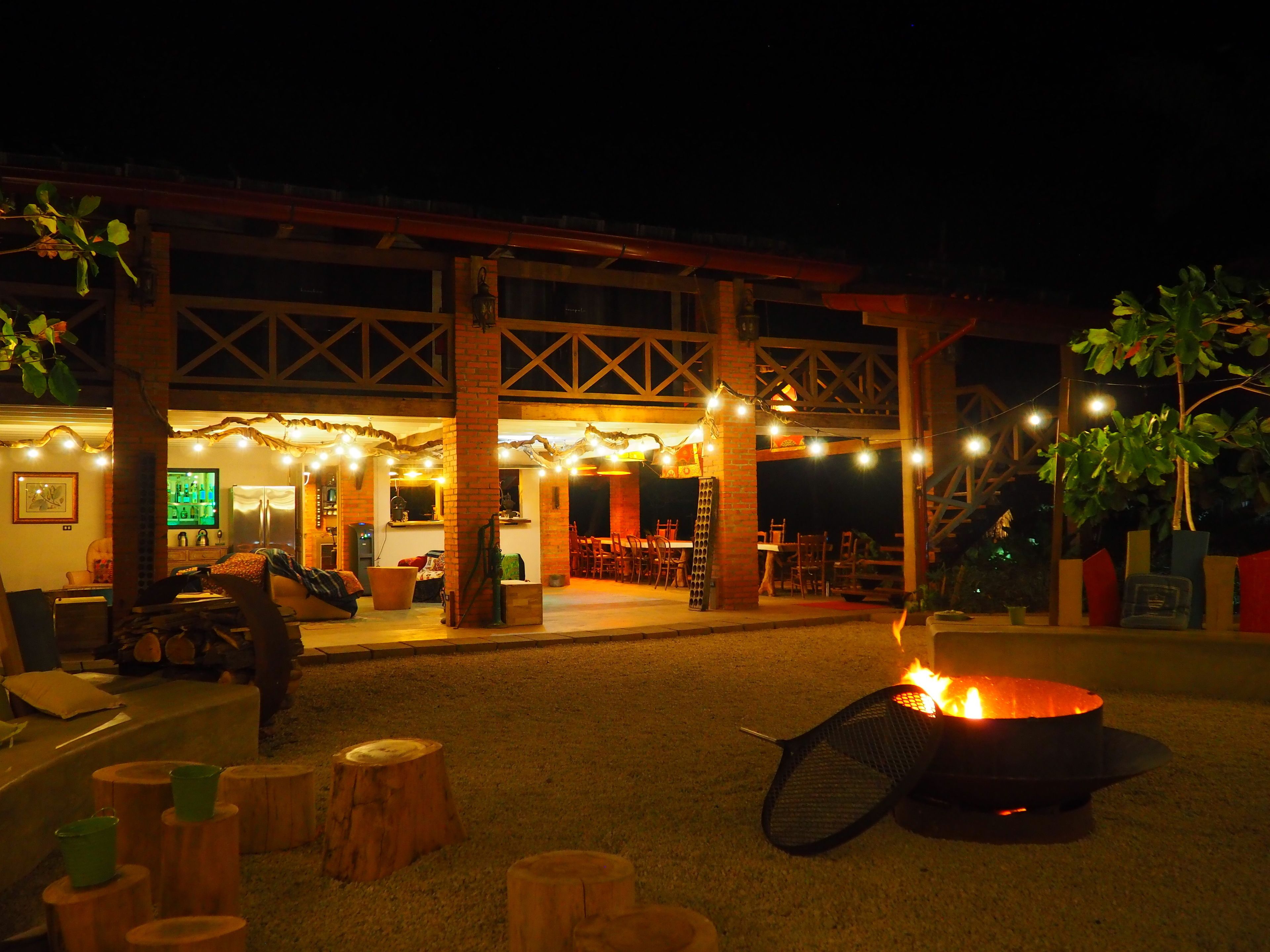 Un espacio al aire libre con una fogata, taburetes de madera y luces colgantes.