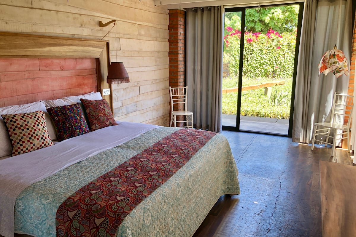 A bedroom with a large bed, a wooden headboard, a hanging lamp, and a view of a garden through a sliding glass door.