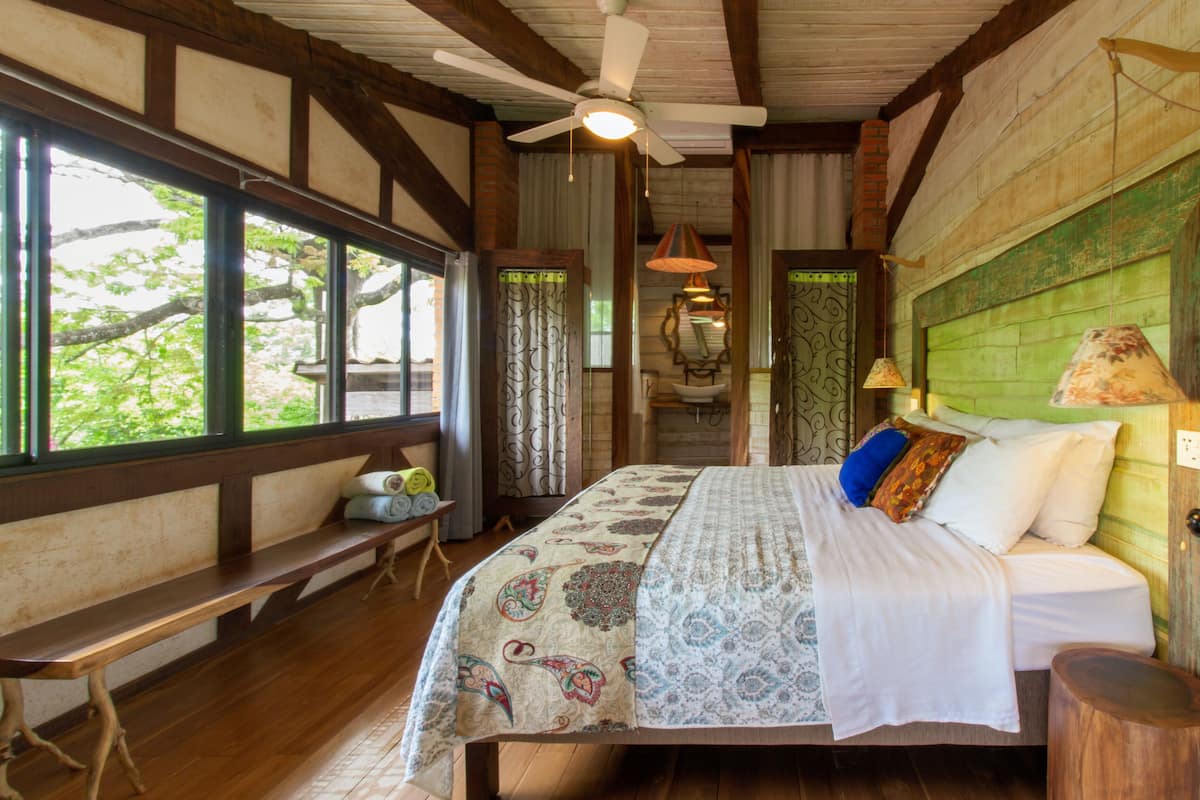 A bedroom with a bed, a bench, a ceiling fan, and large windows.