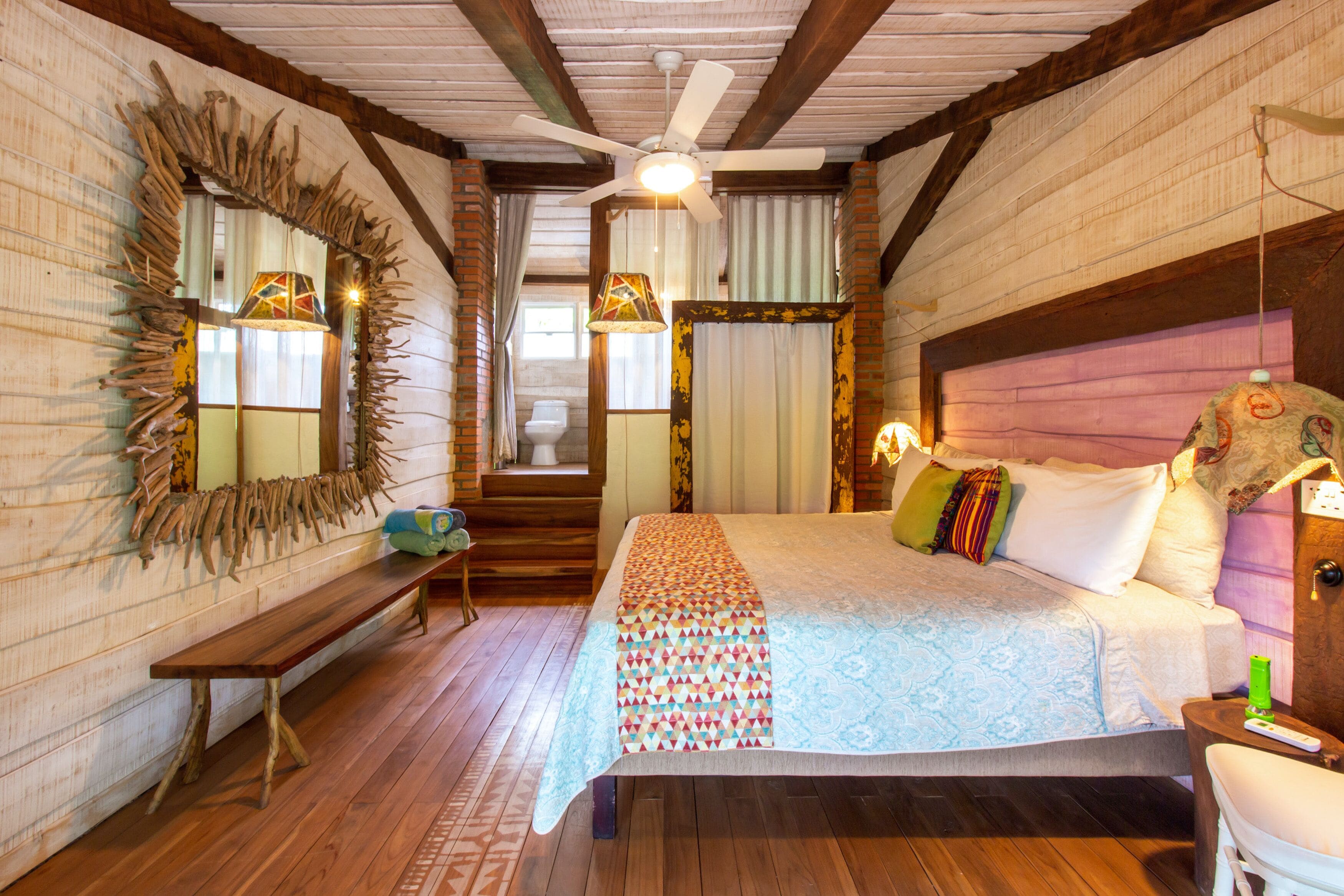 A bedroom with a large bed, a wooden bench, a ceiling fan, and a staircase leading to an upper level.