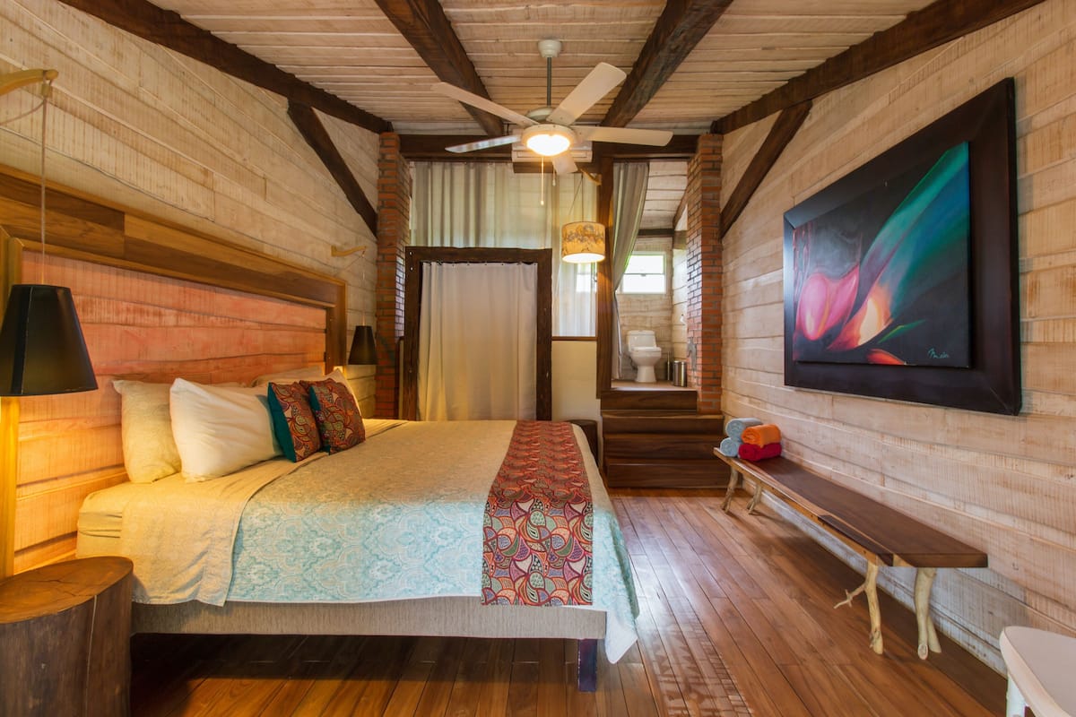 A wooden room with a bed, a bench, a ceiling fan, and a large painting on the wall.