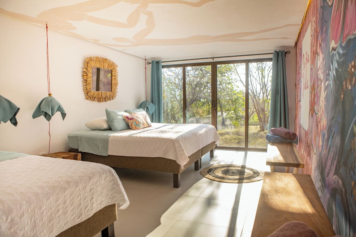 A bedroom with two beds, a large window with curtains, a wooden bench, and a wall painting.