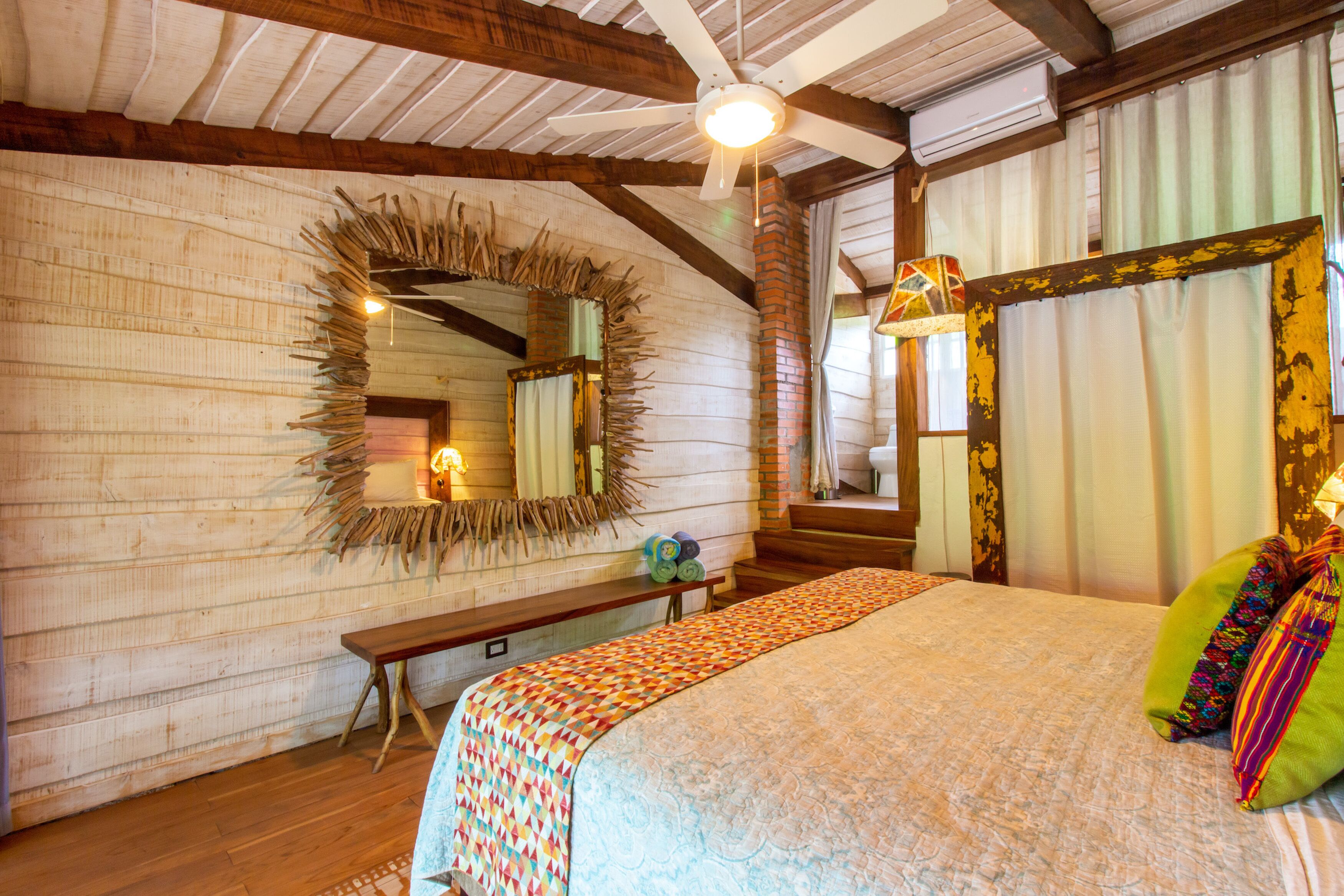 A bedroom with a wooden ceiling, a large bed with a colorful blanket, a bench, a mirror, and a window with curtains.