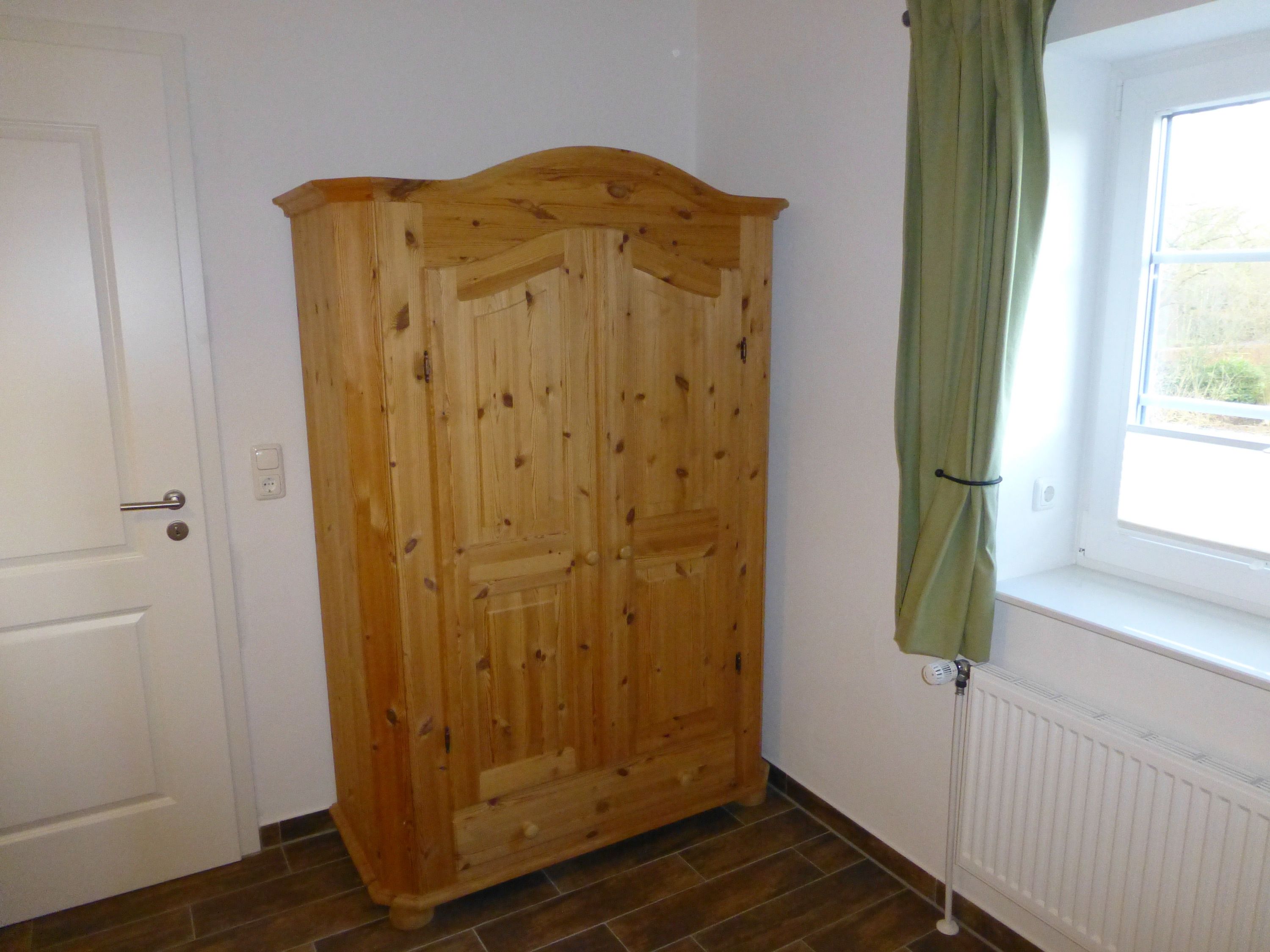 Um guarda-roupa de madeira num quarto com uma porta branca, uma janela com cortina verde e um radiador.
