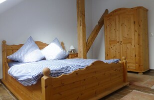 Ein hölzernes Bett mit karierter Bettwäsche, ein Nachttisch mit Lampe und ein Holzschrank in einem Raum mit schragem Dach.