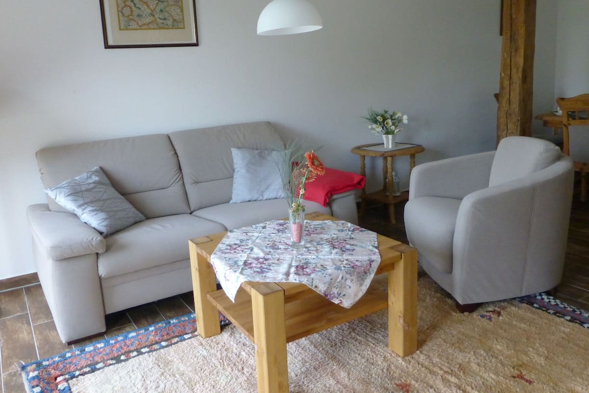 Ein Wohnzimmer mit einer Couch, einem Couchtisch aus Holz mit einer blumigen Tischdecke, einem Sessel, einem kleinen runden Tisch mit Blumen und einer Pendelleuchte.